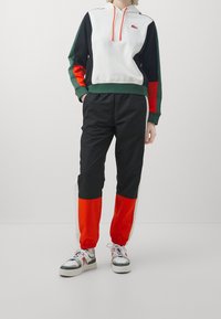 Felpa con cappuccio in bianco, verde, nero e arancione, abbinata a pantaloni da tuta con pannelli neri e arancioni. Presenta polsini elasticizzati e cordino.