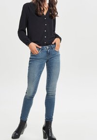 Blusa nera a maniche lunghe con bottoni frontali, abbinata a jeans blu aderenti e stivaletti neri. Tessuto liscio e vestibilità sartoriale.