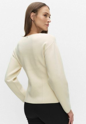 Cremefarvet striktrøje med rund halsudskæring, lange ærmer og diskrete detaljer bagpå. Tekstureret stof med en tætsiddende silhuet.