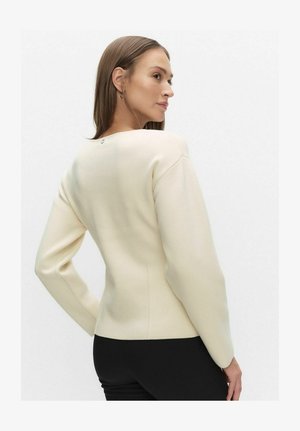 Cremefarvet striktrøje med rund halsudskæring, lange ærmer og diskrete detaljer bagpå. Tekstureret stof med en tætsiddende silhuet.