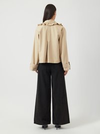 Femme aux longs cheveux noirs portant une veste beige à col à volants et un pantalon noir à jambes larges, debout face à un mur blanc.