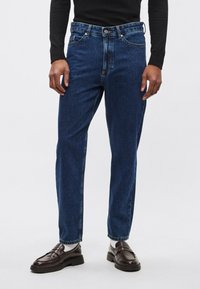 Dark blue denim jeans with a straight-leg design, five pockets, and a button closure at the waist. Smooth texture and classic stitching.