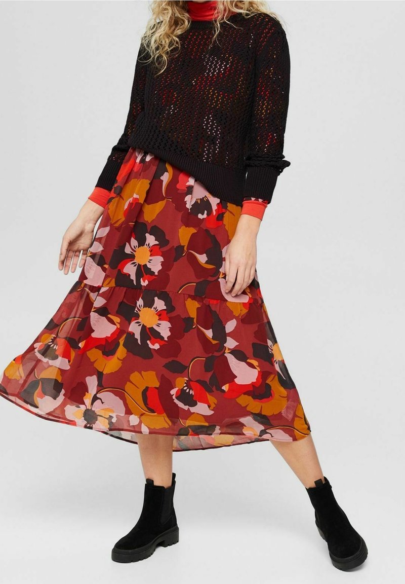 Un pull noir tricoté avec un motif texturé superposé à une jupe maxi à imprimé floral dans des teintes rouges, oranges et roses, associé à des bottines noires.