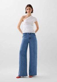 Weißes, einseitig tragbares Oberteil mit sanfter Textur, kombiniert mit hellblauen, weit geschnittenen Jeans. Das Model trägt rote High Heels und hat die Hände in den Taschen.