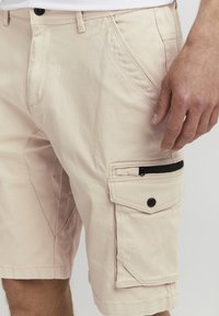 Shorts cargo beiges en coton avec une poche zippée sur le côté, dotés de quincaillerie noire et de détails de couture subtils.