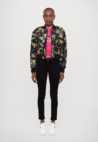 Black bomber jacket with gold and white floral pattern, pink fitted shirt, black skinny jeans, and white sneakers.