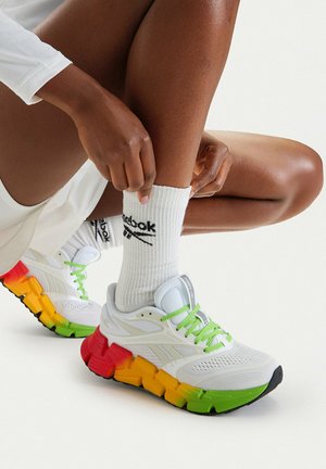 Persona con calcetines blancos de Reebok y zapatillas blancas con una suela multicolor en verde, amarillo y rojo, ajustándose el calcetín mientras está sentada con las piernas dobladas.