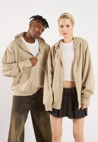 Beige oversized hettegensere med frontglidelås, med store hetter og ribbestrikkede mansjetter. Mørke denimjeans og et svart denimskjørt bæres under.