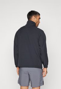 Zwarte lichte jas met een hoge kraag, elastische manchetten en een rechte snit, gedragen over grijze shorts. Gladde stofstructuur.