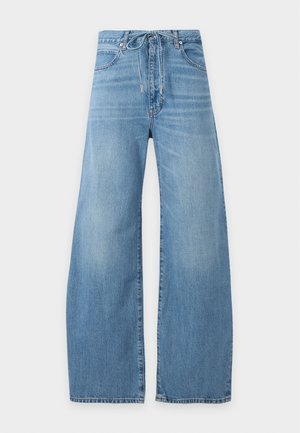 Femme portant une chemise boutonnée jaune pâle, un jean bleu à jambes larges, des sandales noires et tenant un sac à main jaune pâle assorti.