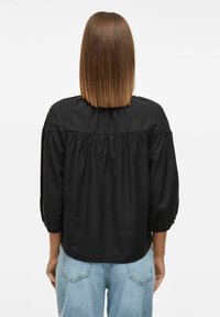 Melna blūze ar apaļu izgriezumu, pūkainām piedurknēm un savilkta aizmugures dizaina. Valkāta ar gaiši zilām augstām džinsām, gluda tekstūra.