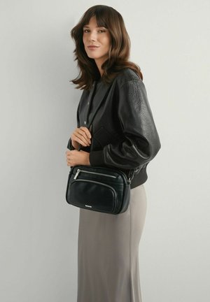 Kobieta o brązowych włosach do ramion, ubrana w czarną skórzaną kurtkę i beżową spódnicę, trzymająca czarną skórzaną torbę na ramię.