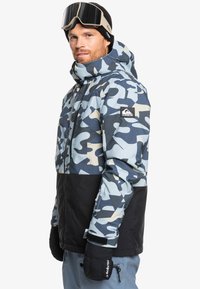 Blå og beige camouflagesnejakke med et sort nederste. Har hætte, lynlåslukning og elastiske manchetter. Indeholder et mærke med logo.