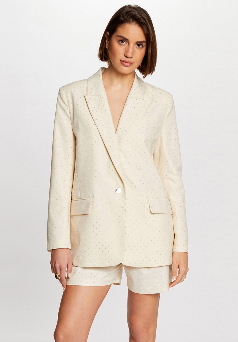 Blazer crema con textura de patrón de diamantes, cierre de un solo botón y dos bolsillos frontales. Lleva puesto con pantalones cortos crema a juego.
