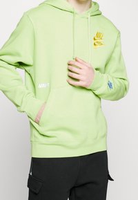 Ljusgrön Nike-huvtröja med framficka, dragsko på huvan, gult logotyp på bröstet och blått logotyp på ärmen. Svarta byxor är synliga.