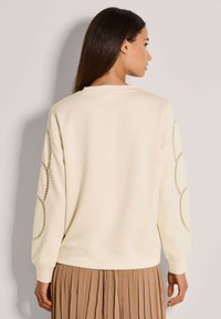 Frau mit langen dunklen Haaren, die einen cremefarbenen Sweatshirt mit runden Ausschnitten an den Ärmeln und einen beigen, plissierten Rock trägt, mit dem Rücken zur Kamera.