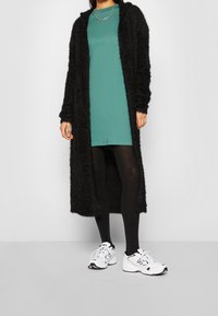 Svart, fluffig lång cardigan över en petrolfärgad klänning, parat med svarta leggings och vita sneakers. Cardiganen har en mjuk, texturerad yta.
