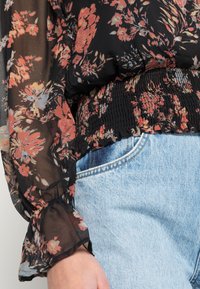 Blusa floral em tecido preto transparente com padrões laranja e azul. Apresenta uma cintura franzida e mangas bufantes. Combinada com jeans de denim claro.