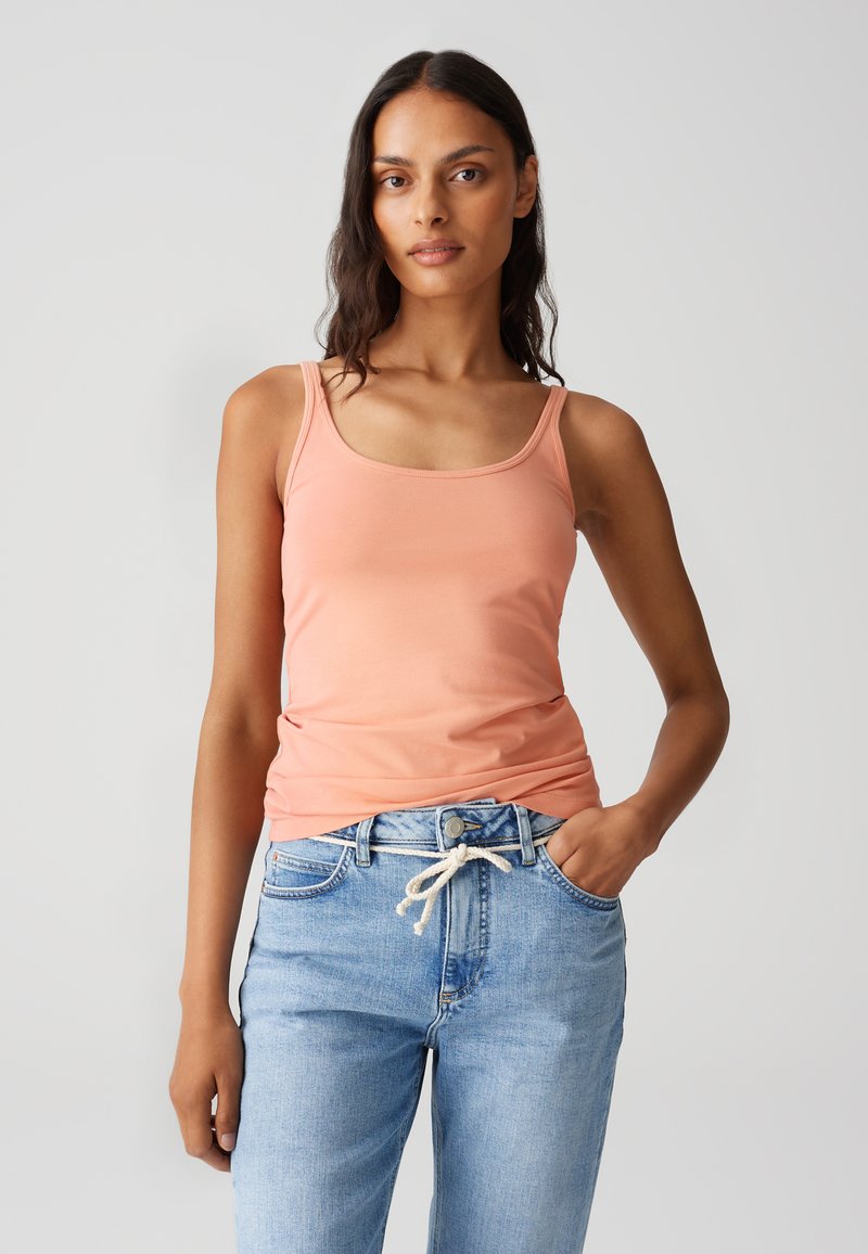 Pfirsichfarbenes Tanktop mit feinen Trägern, aus glattem Stoff gefertigt. Kombiniert mit hellblauen, locker sitzenden Jeans aus Denim mit einer Kordelzugtaillierung.
