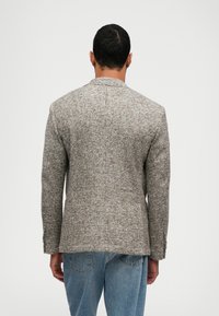 Man wearing a fitted gray plaid blazer and blue jeans, standing facing away against a plain light background.