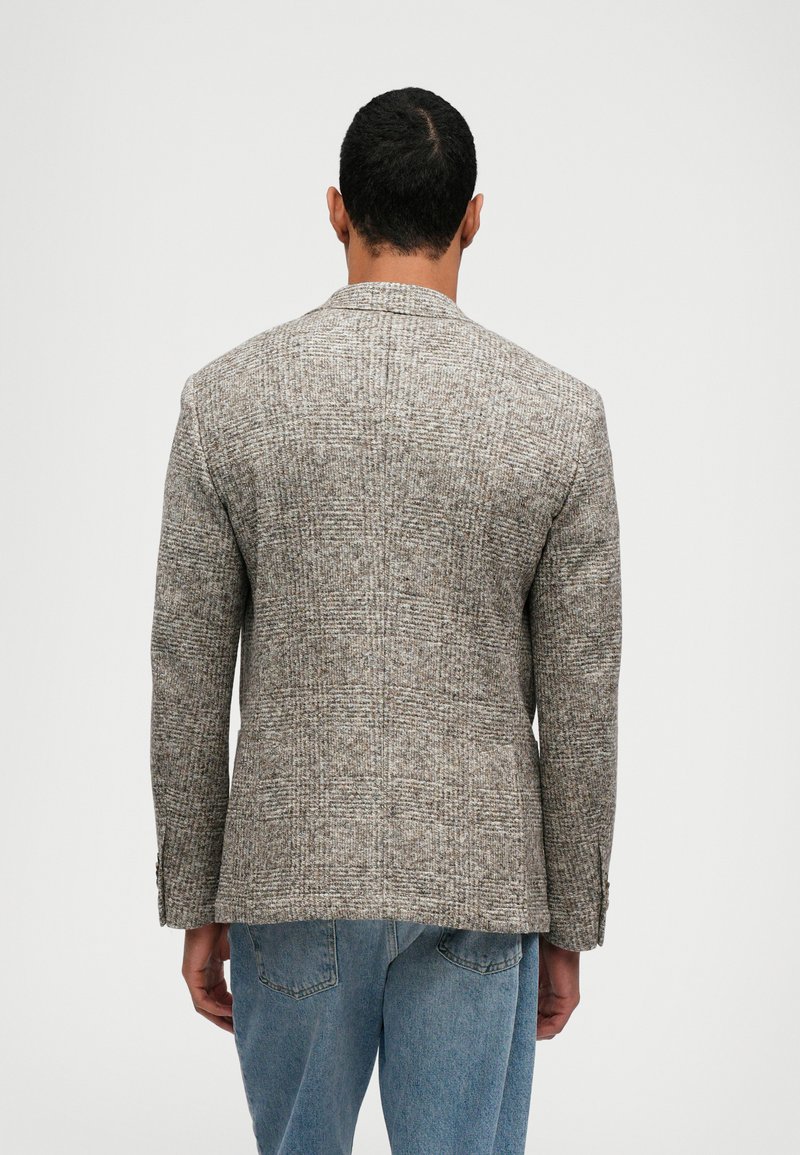 Man in een getailleerd grijs geruit blazer en blauwe jeans, staand met zijn rug naar de camera tegen een effen lichte achtergrond.