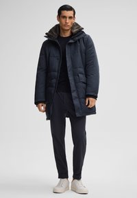 Marineblauwe gewatteerde jas met een nepbontkraag, voorzien van zijzakken, een ritssluiting aan de voorkant en een ontspannen pasvorm, gecombineerd met donkere broeken en witte sneakers.