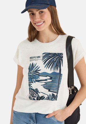 Jeune femme portant un t-shirt blanc avec un imprimé tropical bleu et le texte "SELFCARE", casquette en jean et sac à bandoulière noir, souriante.