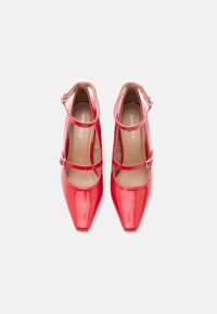 Rode schoenen van lakleer met een puntige neus, twee enkelbandjes en gespsluitingen. Gladde textuur en een gestroomlijnde silhouet.