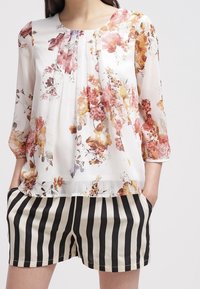 Blusa bianca fluida con motivo floreale rosa, arancione e giallo; presenta un collo rotondo e maniche a tre quarti. Abbinata a pantaloni corti a righe bianche e nere.