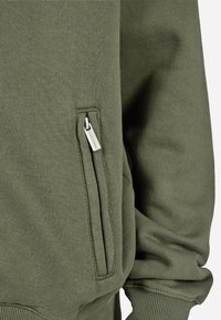 Olīvzaļu džemperis ar teksturētu audumu; tam ir sānu rāvējslēdzēja kabata un ribotie aproces ap malām un piedurknēm.