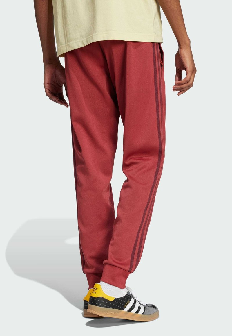 Red athletic pants with three dark stripes along the sides, featuring a tapered design and smooth texture, paired with gray and yellow sneakers.