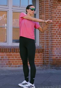 Maglietta atletica rosa a maniche corte, leggings neri e scarpe da ginnastica bianche con accenti neri. Pose di stretching davanti a uno sfondo di muro di mattoni.