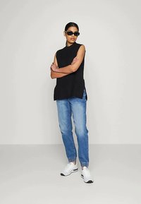 Mulher vestindo uma blusa preta sem mangas, jeans jogger azuis, tênis brancos e óculos de sol pretos, de pé com os braços cruzados contra uma parede branca.