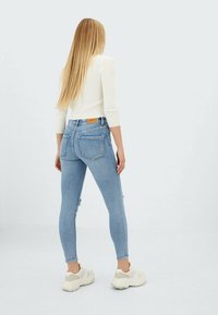 Jeans skinny de color azul claro con detalles desgastados, de cintura alta, y un parche de cuero marrón en la parte trasera. Combinados con zapatillas blancas y una blusa blanca de canalé.