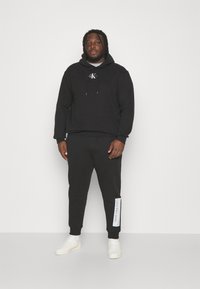 Čierna mikina s kapucňou a logom, čierne tepláky s bielym textom "Calvin Klein Jeans" na boku a biele tenisky. Hladký materiál, voľný strih.