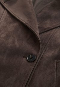 Dunkelbrauner Wildledermantel mit breiten Revers und einem einzelnen schwarzen Vierlochknopf, der nahe der vorderen Mitte befestigt ist.