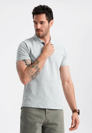 Lys grå polo shirt med korte ærmer, lavet af en bomuldsblanding, med et subtilt logo på brystet. Model i en afslappet positur, tatovering synlig.