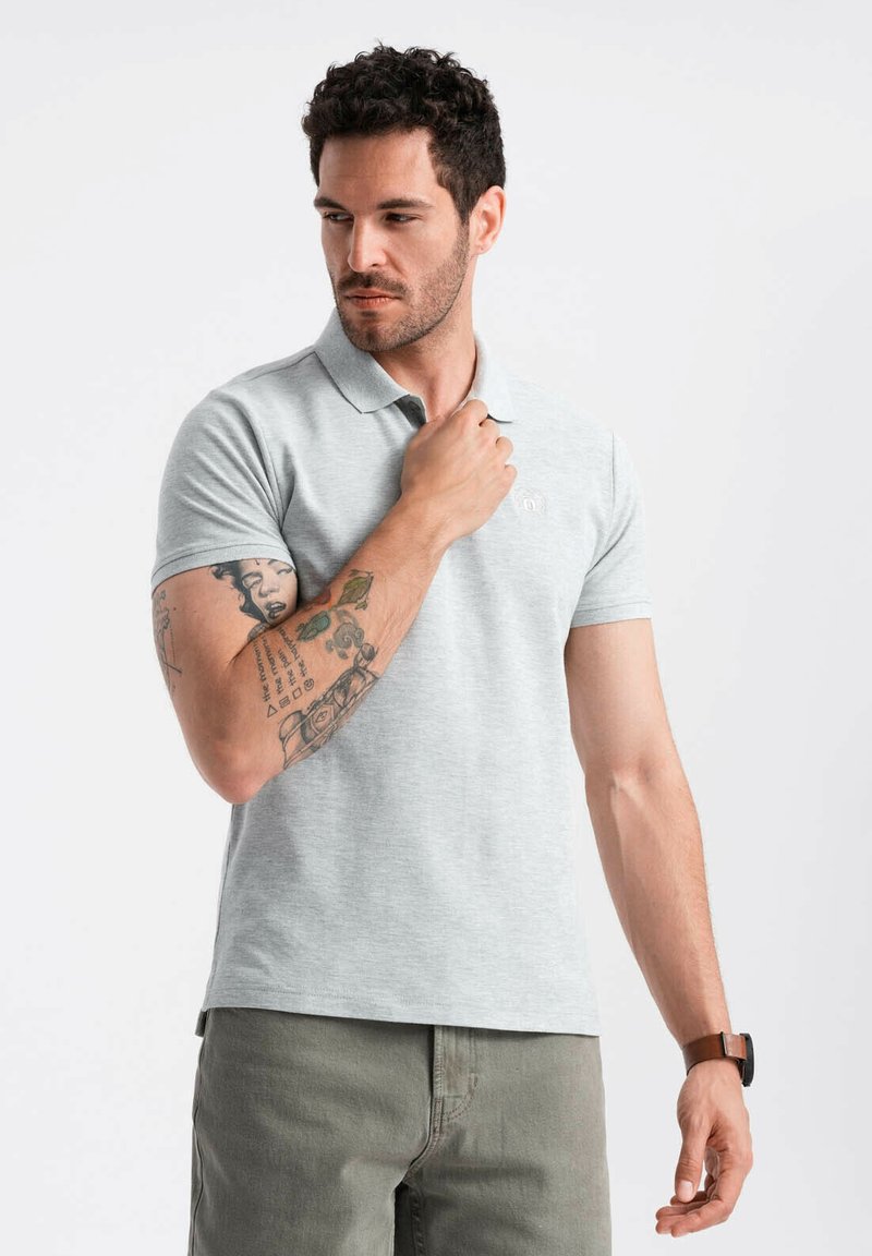 Polo grigio chiaro a maniche corte, realizzato in un misto di cotone, con un logo discreto sul petto. Modello in posa casual, tatuaggio visibile.