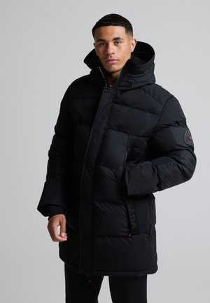 Jonge man die een zwart gewatteerd winterjack met capuchon draagt, staat met één hand gedeeltelijk in de zak, neutrale achtergrond.