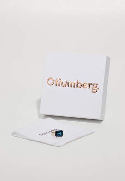 Anillo de gema azul con acentos dorados sobre una tela blanca, con una caja blanca que muestra "Otiumberg" en letras de oro rosa.