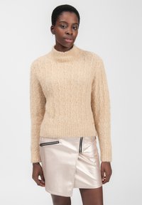 Beige tekstureret striktrøje med høj krave og lange ærmer, parret med en skinnende metallisk pink plisseret nederdel med lynlåsdetalje.