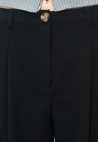 Gros plan sur un pantalon noir avec une fermeture à bouton porté avec un haut zippé gris côtelé.