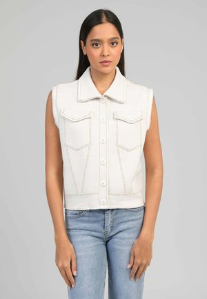 Jeune femme aux longs cheveux bruns portant un gilet blanc sans manches boutonné et un jean bleu clair, debout devant un fond gris uni.