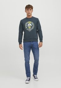 Felpa verde scuro con un logo rotondo che presenta un teschio e rose, abbinata a jeans blu e sneakers navy. Semplice texture a maglia.