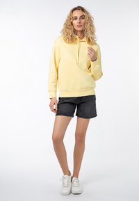 Geel hoodie met trekkoorden, gekoppeld aan zwarte denimshorts. Witte sneakers maken de outfit af. Lichte stof, casual pasvorm.