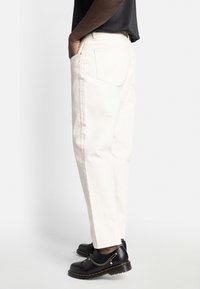 Persoană îmbrăcată în pantaloni albi drepti și pantofi din piele negri cu lanțuri decorative, stând cu mâinile în buzunare.