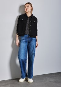 Svart jacka med knappruscher, tillsammans med blå vida denimjeans och vita sneakers. Diskret mönstrad topp under.