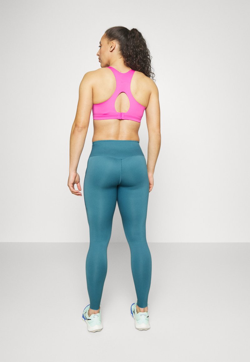 Frau, die mit dem Rücken steht, trägt einen rosa Sport-BH, eine türkisfarbene, hochtaillierte Leggings und weiße Laufschuhe vor einem einfarbigen Hintergrund.