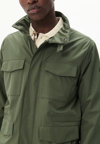 Olivgrüne wasserdichte Jacke mit hohem Kragen, zwei Brusttaschen und zwei Seitentaschen sowie einer glatten, matten Textur.
