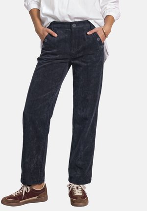 Personne portant un pantalon en velours côtelé bleu foncé avec des boutons sur les poches et des baskets marron avec des lacets blancs, mains dans les poches, chemise blanche.